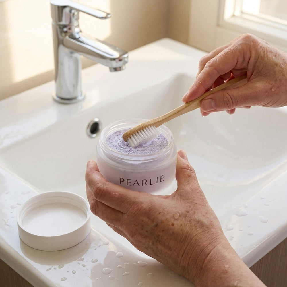 A wet bamboo toothbrush dipped into the open Pearlie jar at the bathroom sink